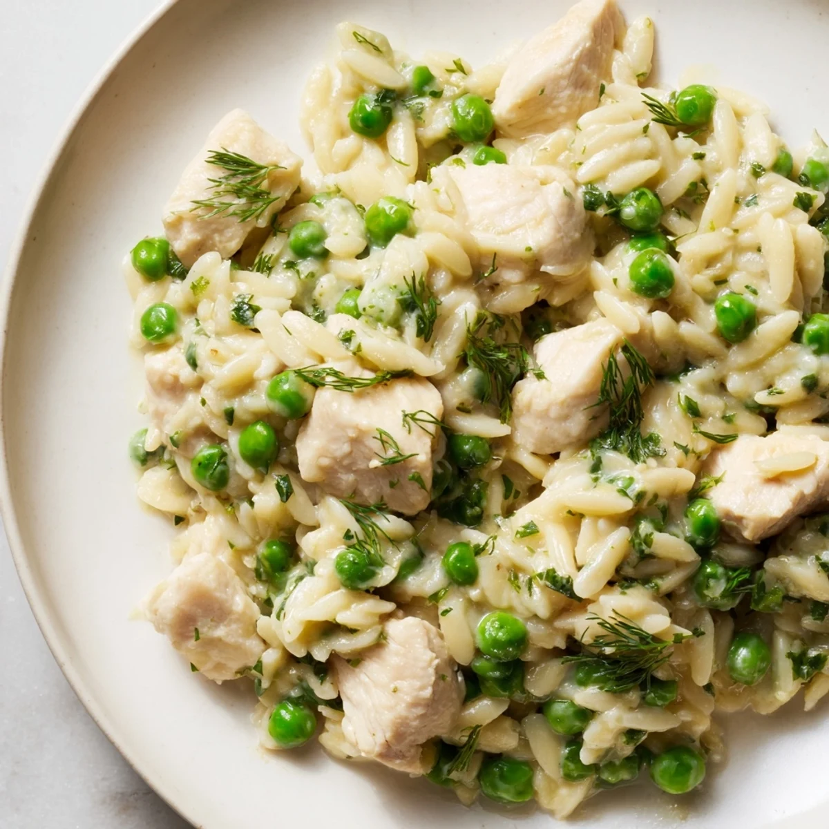 Golden-brown chicken pieces simmer with orzo pasta, green peas, and fresh herbs in a bright lemon sauce inside a hot skillet.