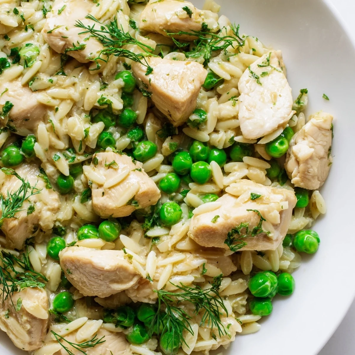 A close-up view of Lemon Herb Chicken Orzo Skillet shows tender chicken nestled in creamy orzo with vibrant green peas and herbs.