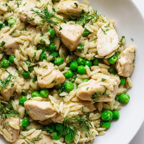 A close-up view of Lemon Herb Chicken Orzo Skillet shows tender chicken nestled in creamy orzo with vibrant green peas and herbs.